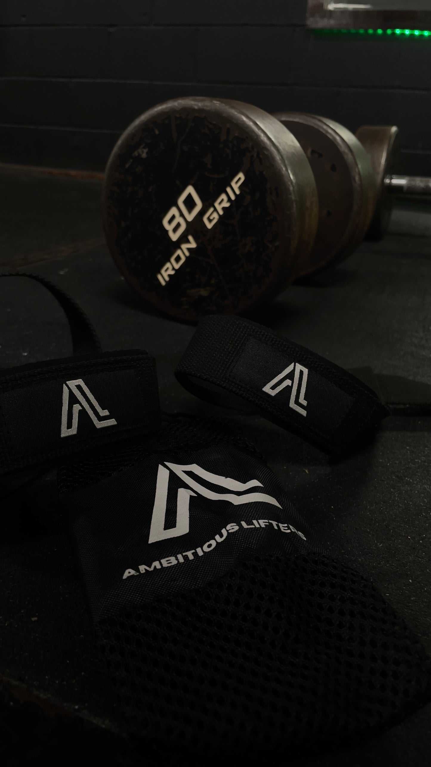 Dumbbell with '80 Iron Grip' label and black wrist wraps with 'Ambitious Lifter' logo on a dark surface.
