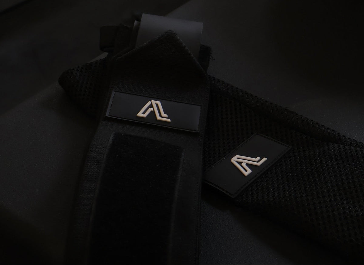 Black weightlifting wrist straps with a gold stylized 'A' logo, shown in a dimly lit gym setting.